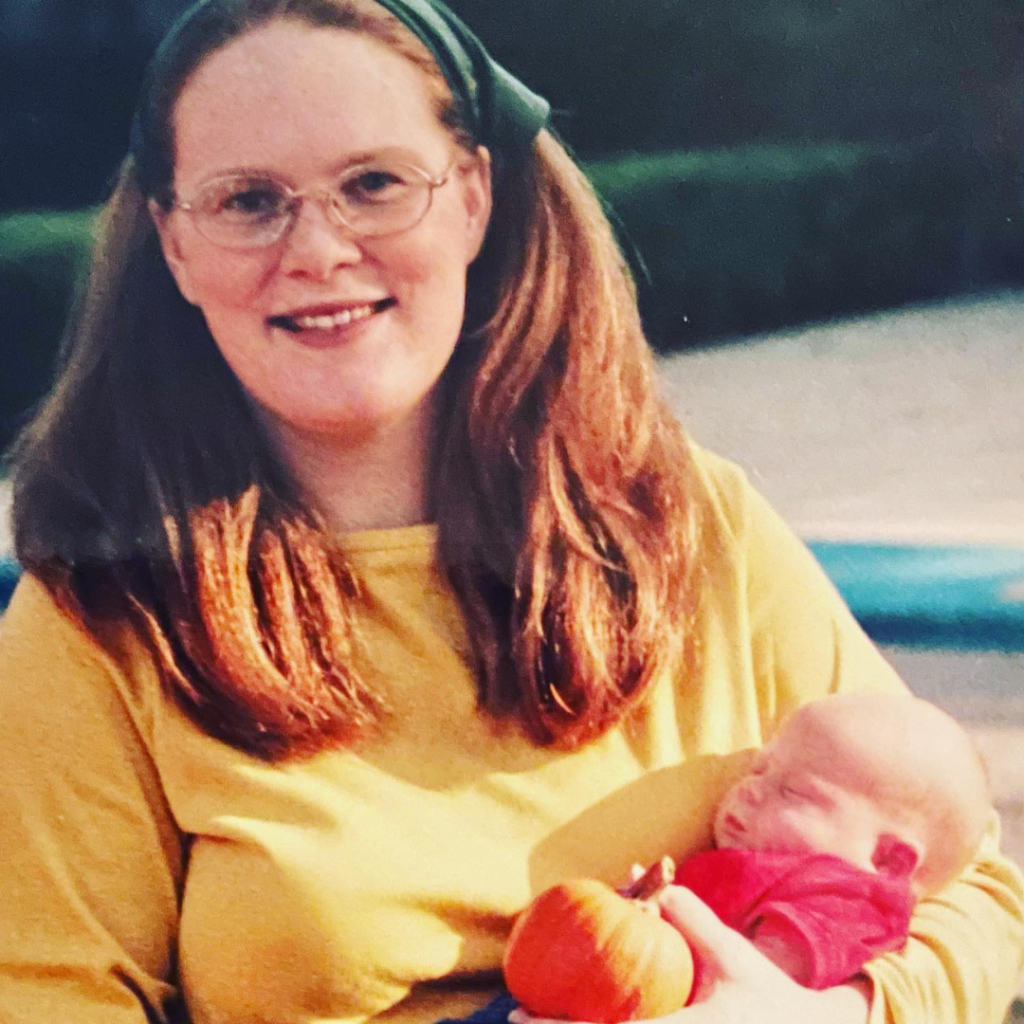 a young fundamentalist woman with red hair and green head covering holds a baby in a red shirt